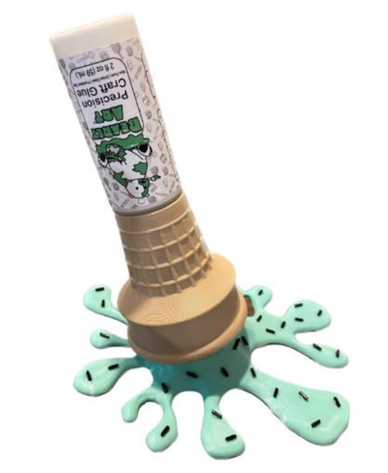 Może przedstawiać: Nowatorski klej w sztyfcie z białą nasadką i etykietą z napisem "Beardy Art Precision Craft Glue 59 mL". Klej w sztyfcie umieszczony jest w brązowym rożku do lod&oacute;w, kt&oacute;ry umieszczony jest na jasnoniebieskiej, stopionej bazie przypominającej lody z czarnymi posypkami.