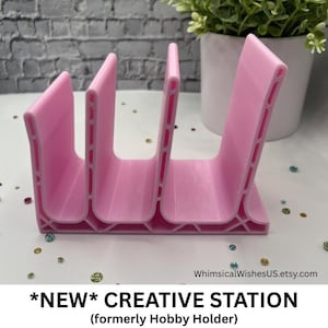 May include: A light pink plastic organizer with three vertical slots for holding items. The structure features a reinforced base with a grid-like pattern and open sides. The text "*NEW* CREATIVE STATION (formerly Hobby Holder)" is printed below the object.