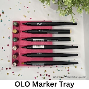 May include: A bright pink rectangular plastic tray with six horizontal slots, each holding a black OLO marker. The tray features six circular indentations on the left side, each containing a marker cap labeled with color codes like YG 2.7 and OR 7.0.