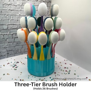 May include: A three-tier brush holder in turquoise holds 26 makeup brushes. The brushes have white oval heads and colorful handles in shades of orange, yellow, pink, green, and purple. The text at the bottom reads "Three-Tier Brush Holder (Holds 26 Brushes)."