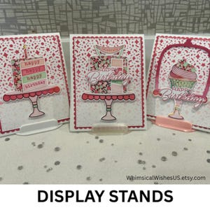 May include: Three birthday-themed display stands. Each stand features a cake or cupcake design with the words "Happy Birthday." The stands are white with red scalloped edges and a clear plastic base. The text "DISPLAY STANDS" is at the bottom.