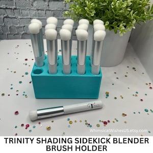 May include: A teal brush holder with multiple silver and white makeup brushes. The holder is rectangular with several holes to hold the brushes upright. One brush lies in front of the holder. The text "TRINITY SHADING SIDEKICK BLENDER BRUSH HOLDER" is at the bottom.