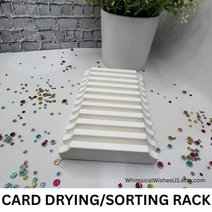 May include: A white card drying and sorting rack with multiple horizontal slots. The rack is on a white surface with colorful sequins scattered around. A white pot with a green plant is in the background. Text at the bottom reads "CARD DRYING/SORTING RACK".