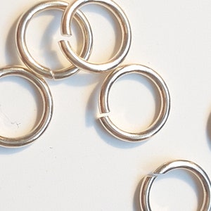 May include: Six silver jump rings, also known as split rings, are shown on a white surface. These rings are used for attaching charms, pendants, or other jewelry components.