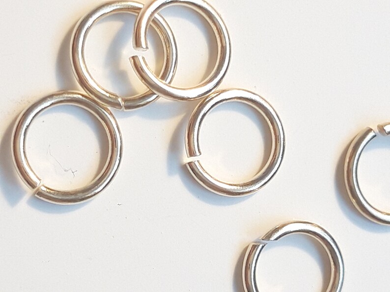 May include: Six silver jump rings, also known as split rings, are shown on a white surface. These rings are used for attaching charms, pendants, or other jewelry components.
