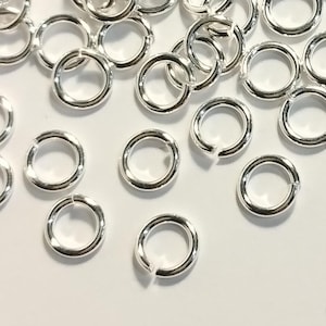 May include: A selection of small, shiny, silver jump rings scattered on a white surface. The jump rings are open, with a small gap in the circle, suitable for jewellery making or other crafts.