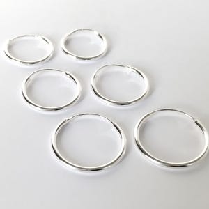 May include: Six silver hoop earrings are displayed on a white surface. The earrings are circular, with a smooth, polished finish. The light reflects off the metal, creating highlights and shadows. The earrings are of varying sizes.