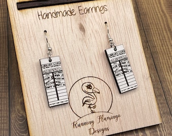 Puede incluir: Pendientes de madera hechos a mano con forma de tarjetas de biblioteca con el texto "LIBRARY CARD" en ellos. Los pendientes están en un expositor de madera con el texto "Running Flamingos Designs".