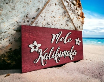 May include: A red wooden sign with the text "Mele Kalikimaka" in white lettering. The sign is decorated with white flower cutouts and is hanging from a rope.