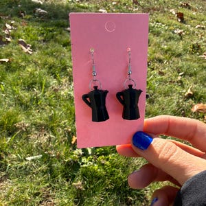 May include: Black coffee pot earrings with silver-colored hooks, displayed on a pink card. The earrings are held in front of a grassy background. The earrings are a fun accessory for coffee lovers.