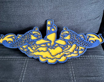 May include: A blue and gold wooden wall decor featuring a stylized submarine with dolphins on either side. The submarine is in the center of the design and is surrounded by waves.