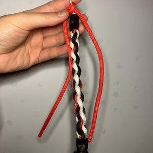 May include: A handmade keychain with a braided cord in red, black, and white. The braided section is attached to a black, textured base and a red cord loop with a metal ring.