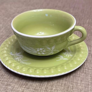 以下が含まれることがあります： 明るい緑色のティーカップとソーサーのセット。カップには取っ手と白い花の模様があります。ソーサーはマッチしたデザインとビーズの縁があります。セットはセラミック製で、白い縁があります。