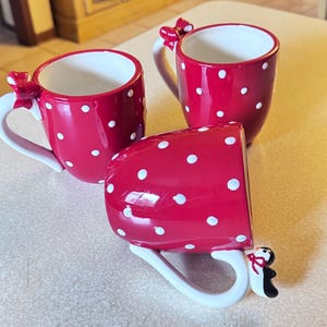 May include: Three red ceramic mugs with white polka dots. Each mug has a white handle with a bow detail and a penguin accent. The mugs are arranged on a light-colored surface.