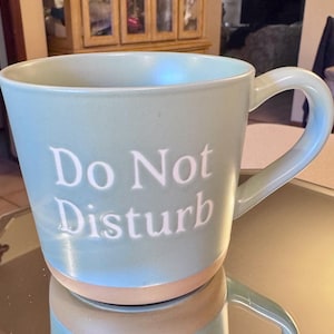 May include: A light blue ceramic mug with the words "Do Not Disturb" in white lettering. The mug has a rounded handle and a tan-colored base. The mug is sitting on a reflective surface.