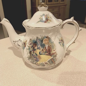 Op de afbeelding: Een witte theepot met een gouden rand en bloemenpatroon. De theepot heeft een geschilderd tafereel van een stel dat in een tuin danst.