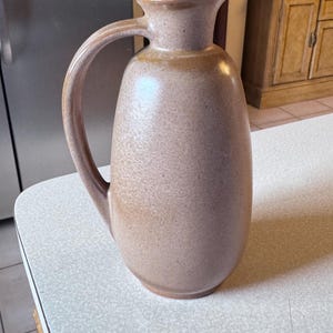 May include: A tan ceramic jug with a large handle and a cork stopper. The jug has a rounded body and a spout for pouring. The surface has a speckled texture. The jug is on a white countertop.