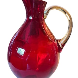 Puede incluir: Una jarra de cristal rojo rubí vibrante con cuerpo bulboso, cuello estrecho con pico vertedor ensanchado y un asa curva de cristal color ámbar translúcido unida al lateral. La superficie del cristal es lisa y tiene un acabado reflectante.