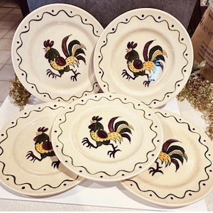 Puede incluir: Un juego de cuatro platos redondos de cerámica con una base de color crema moteado. Cada plato presenta una ilustración central de un gallo con plumas amarillas, verdes y rojo oscuro, rodeado por un borde de línea ondulada verde oscuro.