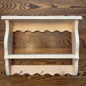 May include: A light-colored wooden wall shelf with a scalloped edge and three pegs below. The shelf has a top shelf and a lower shelf, with curved side supports. The wood grain is visible.