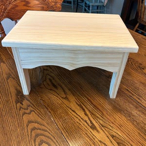 May include: A small, rectangular wooden stool made of light-colored, unfinished pine. The stool features a flat top surface with a visible wood grain pattern and four sturdy legs. The front apron of the stool has a decorative arched cutout design.