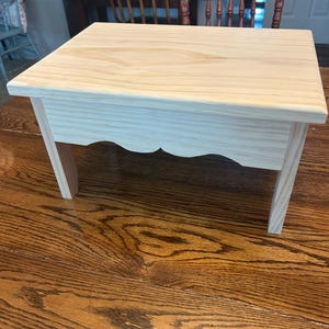 May include: A small, rectangular wooden stool made of light-colored, unfinished pine. The stool features a flat top surface, four straight legs, and a decorative scalloped apron detail along the front edge, showcasing the natural wood grain.