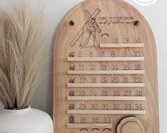 May include: An arched wooden Lenten countdown calendar featuring an engraved illustration of a figure carrying a cross, the text "TAKE UP YOUR CROSS and follow me," and numbered slots from 1 to 40, with a circular wooden piece and a logo at the top.