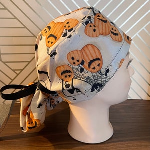 Puede incluir: Un gorro de quirófano blanco con una cinta negra, con un estampado de Halloween de calabazas naranjas con ojos y bocas negros. Las calabazas están dispuestas en un patrón sobre un fondo blanco.