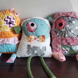 May include: Three handmade plush toys with different patterns and colors. The first toy is orange with a yellow and purple eye, the second is white with a blue and gray eye, and the third is pink with a red and gray eye.