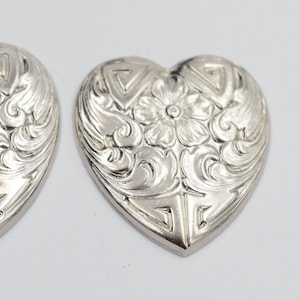 Puede incluir: Dos encantos de plata en forma de corazón con un intrincado diseño floral.