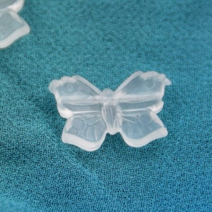 May include: Two clear plastic butterfly-shaped charms on a teal fabric background.