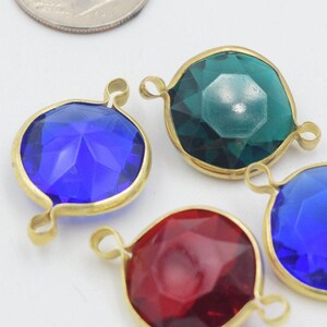 May include: Four colorful gemstone pendants with gold-colored metal settings. The gemstones are blue, green, and red. Each pendant has a loop at the top for attaching to a chain or other jewelry component. The pendants are on a white surface.
