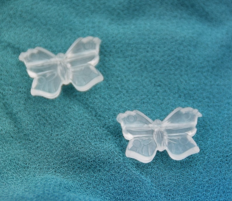 May include: Two clear plastic butterfly-shaped charms on a teal fabric background.