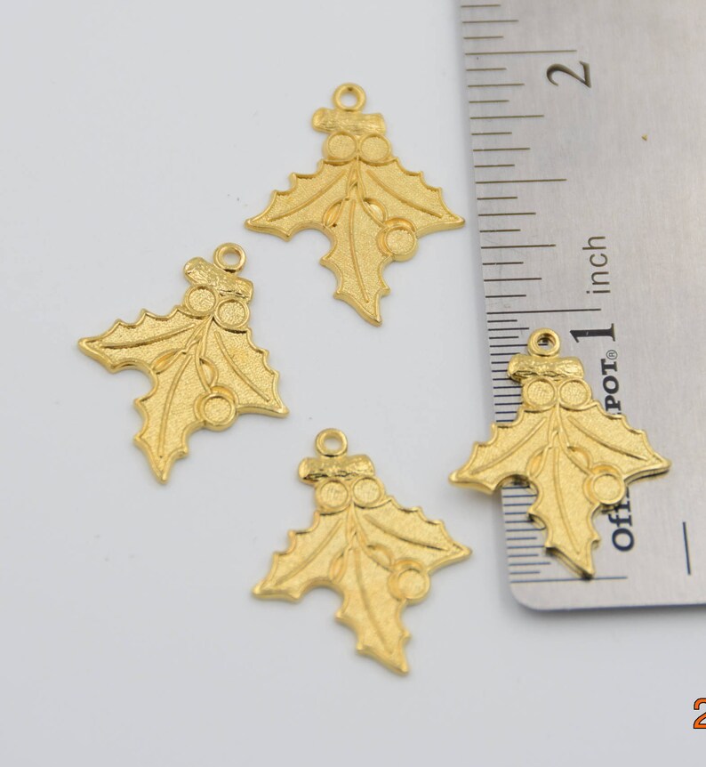 May include: Four gold-colored holly leaf charms with red berries. The charms are approximately 1 inch in size and are shown next to a ruler.