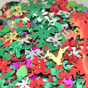 May include: A close-up of a collection of colorful confetti in various shapes, including snowflakes, stars, and floral designs. The confetti is primarily red, green, silver, gold, and purple. The confetti is scattered and overlapping, creating a festive appearance.