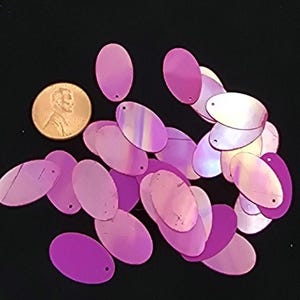 May include: A collection of iridescent, oval-shaped sequins in shades of pink and purple, scattered on a black surface. A U.S. penny is included for size comparison. Each sequin has a small hole for sewing or stringing.