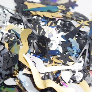 May include: A collection of assorted sequins in various shapes and colors. The sequins include stars, flowers, and abstract shapes in black, silver, gold, and iridescent blue. These decorative embellishments are ideal for crafts and sewing projects.