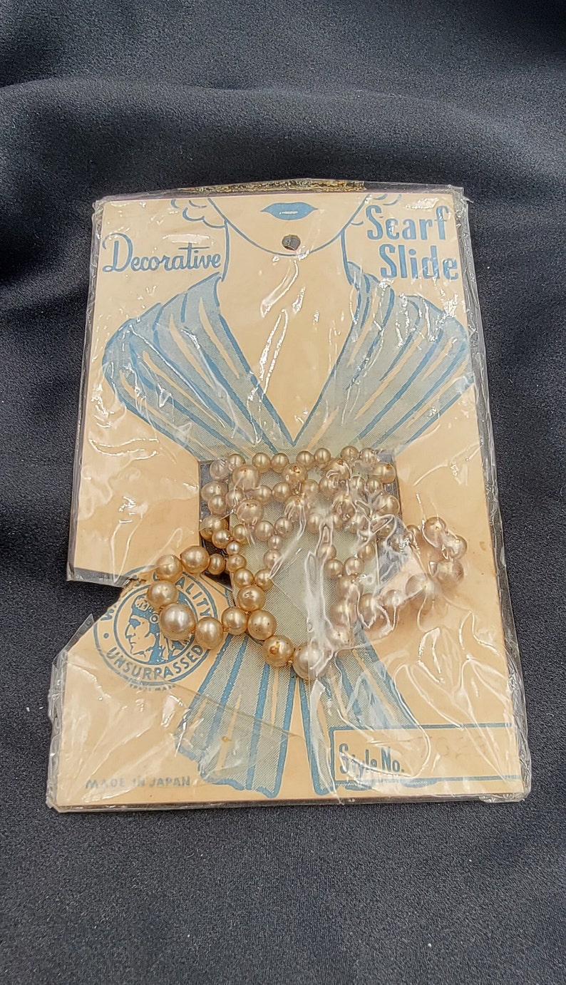 May include: A vintage package containing a decorative scarf slide. The slide is a string of faux pearls in a light brown color. The package is made in Japan and features the text "Scarf Slide" and "Decorative".