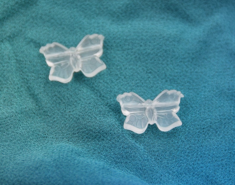 May include: Two clear plastic butterfly-shaped charms on a teal fabric background.