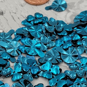 May include: A large pile of teal blue metallic flower-shaped sequins. The sequins are scattered on a white surface. A penny is in the background for scale.