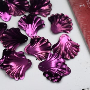 May include: A collection of purple iridescent sequins shaped like seashells. The sequins have a metallic finish and are approximately 1 inch in size.