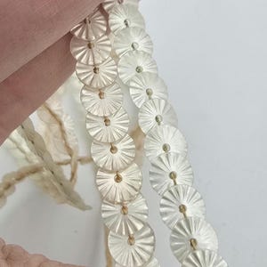 Puede incluir: Dos hebras de lentejuelas blancas iridiscentes con borde festoneado. Las lentejuelas están enhebradas en un hilo fino dorado.