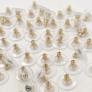 May include: A large group of clear plastic earring backs with gold metal posts. The backs are designed to be used with stud earrings.