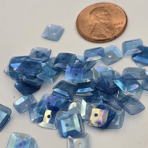 May include: A pile of iridescent blue square sequins with a hole in the center. The sequins are scattered on a white surface. A penny is in the background for size reference.