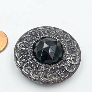 May include: A silver button with a black faceted glass center. The button has a decorative design around the edge.