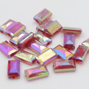 Puede incluir: Un grupo de cuentas rectangulares de vidrio iridiscente rosa y dorado. Las cuentas tienen un diseño facetado y están dispersas sobre una superficie blanca.