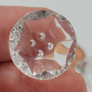 May include: Clear glass button with a star-shaped design and four holes. The button has a textured surface.