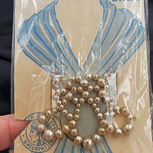 May include: A vintage package of pearl scarf slides, featuring a woman's illustration wearing a scarf. The package is labeled "Decorative Scarf Slide" and "Unsurpassed" with a logo of a Native American head. The package also says "Style No. 752" and "Made in Japan".