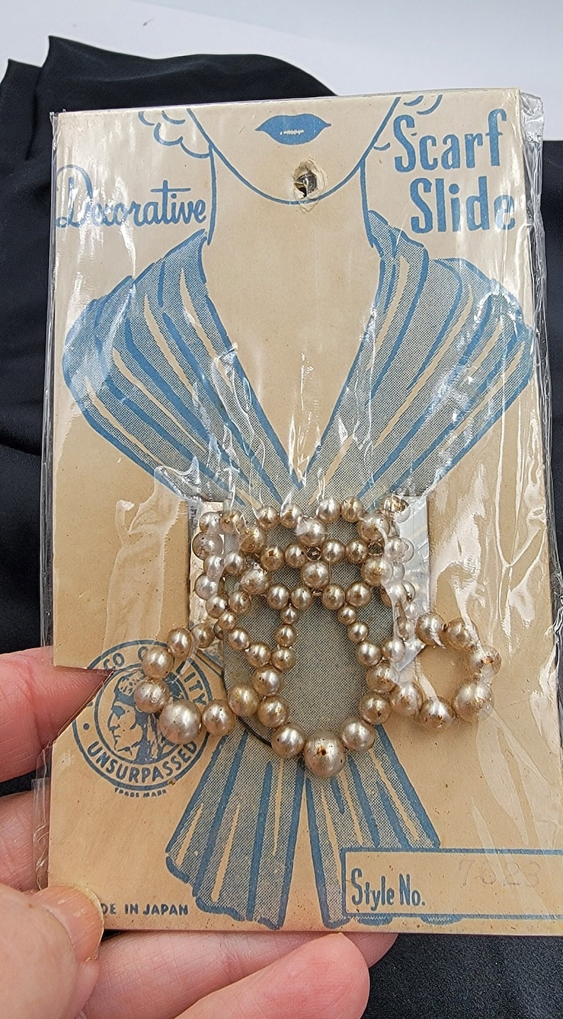 May include: A vintage package of pearl scarf slides, featuring a woman's illustration wearing a scarf. The package is labeled "Decorative Scarf Slide" and "Unsurpassed" with a logo of a Native American head. The package also says "Style No. 752" and "Made in Japan".