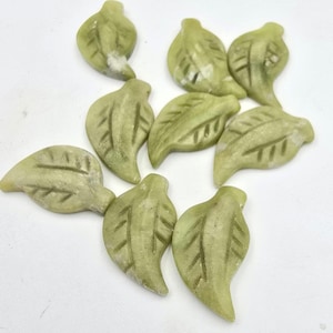 May include: A scattering of small, pale green leaf-shaped stones. Each stone features carved details depicting leaf veins. The leaves are arranged on a white surface, highlighting their natural, earthy hues. These decorative stones could be used for crafts or home decor.